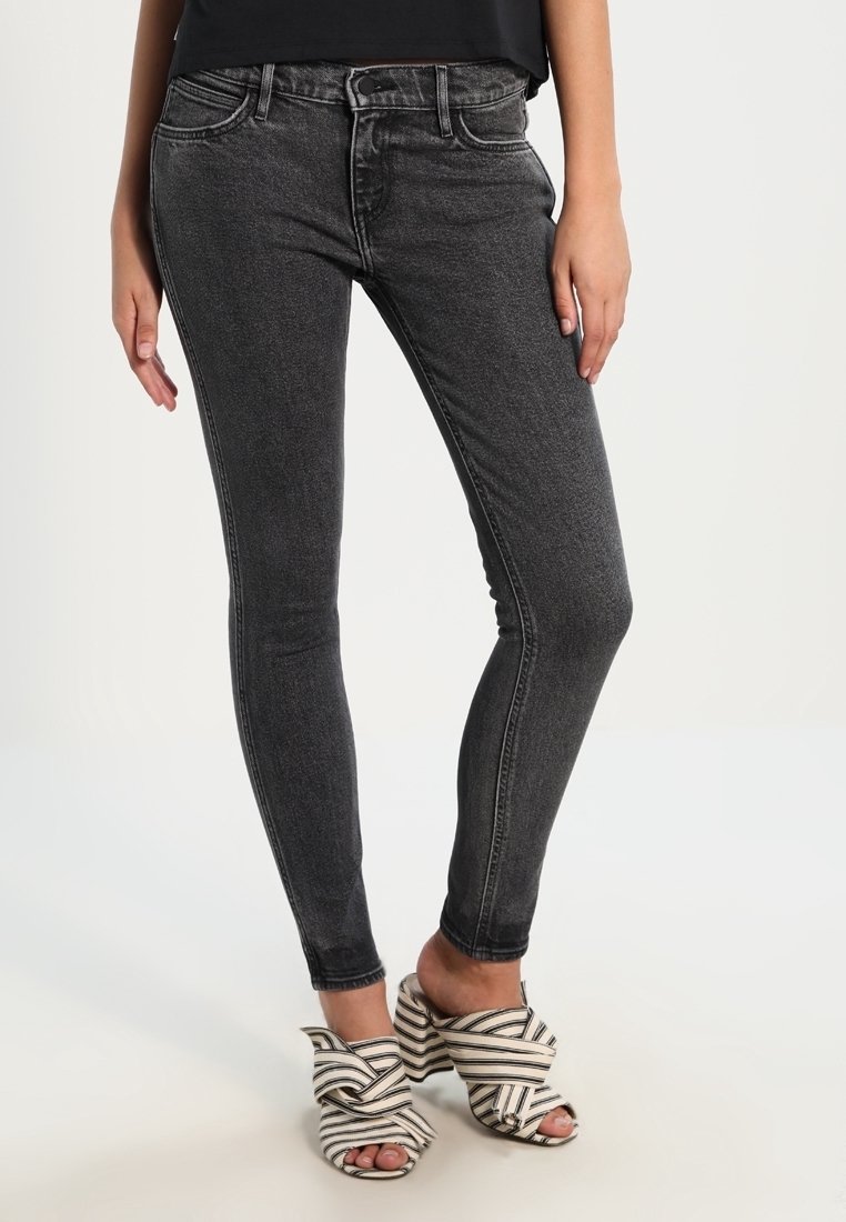 Personne portant un jean skinny gris foncé assorti à des sandales à talons ouvertes à rayures blanches et noires, debout sur un fond clair uni.