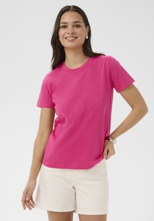Femme souriante, portant un t-shirt rose vif à manches courtes et un short blanc, debout avec une main sur la hanche devant un fond uni.