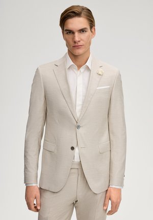 Uomo con completo beige, camicia bianca e spilla floreale sul rever, in piedi davanti a uno sfondo grigio uniforme.