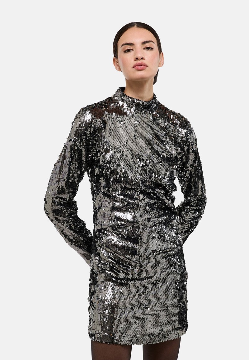 Robe pailletée avec col montant, manches longues et silhouette moulante. Présente des paillettes argentées et noires, créant une surface brillante et texturée.
