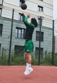 En basketspelare i en grön och svart tröja samt gröna shorts hoppar medan han skjuter en svart basketboll, iförd vita sneakers och ankelsockor.