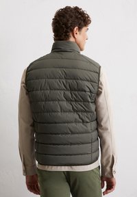 Gepolsterte ärmellose Weste in Dunkelgrün mit gestepptem Design. Der Kragen ist hoch, darunter befindet sich ein beige langärmliges Hemd.