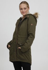 Alyvuogių žalios spalvos parka su kailiniu pamušalu, priekyje zip užtrauktukas, du šoniniai kišeniai ir mygtukų akcentai. Audinys struktūrizuotas, su pritaikytu siluetu.