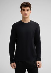 Schwarz langärmliges Rundhals-Shirt aus strukturiertem Stoff. Einfache Gestaltung, minimale Nähte und eine entspannte Passform, kombiniert mit dunklen Hosen.