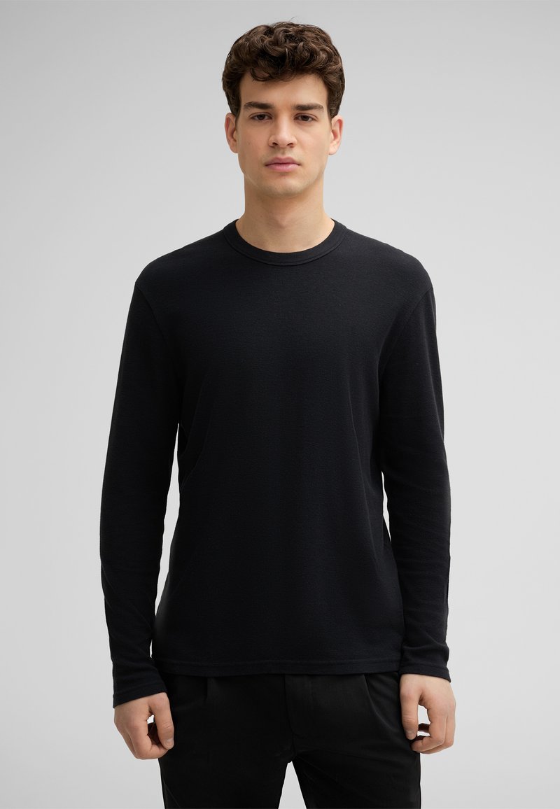 Schwarz langärmliges Rundhals-Shirt aus strukturiertem Stoff. Einfache Gestaltung, minimale Nähte und eine entspannte Passform, kombiniert mit dunklen Hosen.