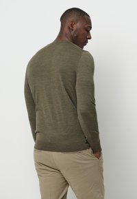 Jersey de color verde oliva de manga larga con cuello redondo y puños acanalados, combinado con pantalones beige claros, visto desde la parte posterior.