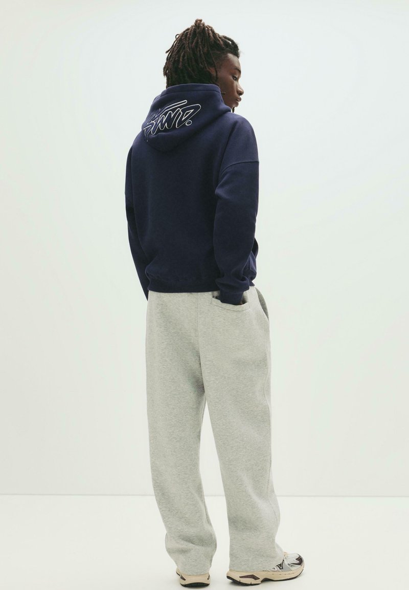 Navy hooded sweatshirt with white logo on the back, paired with light gray sweatpants. The outfit features a relaxed fit and soft fabric.