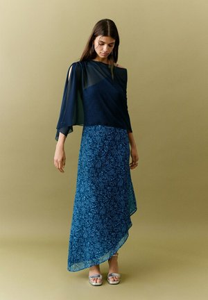 Ženska, oblečena v temnomodno modro enoročno opravo z ohlapnim rokavom in modro cvetlično asimetrično krilo, stoji pred enobarvnim ozadjem.