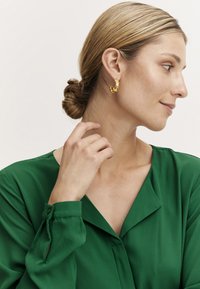Gold statement earrings with a unique design, worn by a person in a green blouse. The earrings have intricate details and a textured finish.