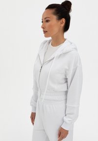 Felpa bianca con zip realizzata in tessuto morbido, con vita e polsini elasticizzati, dotata di tasca a marsupio e cappuccio regolabile con cordino.