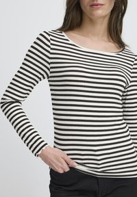 Femeie îmbrăcată cu un tricou cu dungi negre și albe, cu mâneci lungi și decolteu larg, cu mâna odihnind în buzunarul pantalonilor negri.