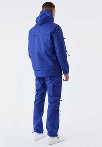 Blauwe waterdichte jas met capuchon, elastische manchetten en verstelbare zoom, gecombineerd met bijpassende blauwe cargobroek. Witte sneakers maken de look compleet.