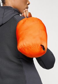 Oranžs iepakojums, izgatavots no viegla materiāla, ar režģa rakstu, savilktu augšu un mazu melnu akcentu. Valkāts uz pleca pret melnu apģērbu.