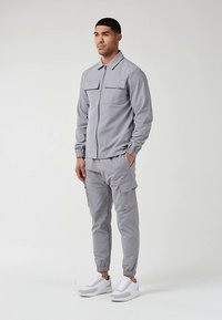 Uomo in piedi, che indossa una giacca leggera grigia con zip e pantaloni cargo, con sneakers bianche, mani nelle tasche, sfondo neutro.