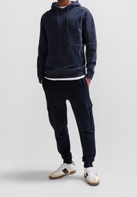 Marinblå sweatshirt med huva och framficka, ihop med matchande joggers med cargofickor. Bärs med vita sneakers och bruna accenter.