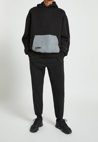 Svart huvtröja med en grå ficka, med en logotyp. Bärs med svarta avsmalnande byxor och svarta träningsskor. Minimalistisk design och löst snitt.