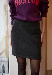 Grijze wollen kokerrok met achterwaartse plooien en zijzakken, gecombineerd met een paarse sweater met rode tekst "BOSTON" en 1980.