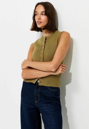 Vrouw met kort bruin haar die een mouwloze olijfgroene top met knopen en een donkerblauwe spijkerbroek draagt, staand met gekruiste armen tegen een witte muur.