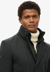 Ciemnoszary płaszcz z wełny z domieszką, w wzór w jodełkę, z czarną kurtką z kołnierzem pod spodem oraz kremowym prążkowanym golfem.