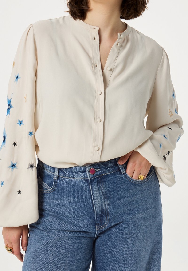 Femme portant un chemisier crème avec broderie d'étoiles bleues sur les manches et un jean bleu taille haute, main dans la poche.