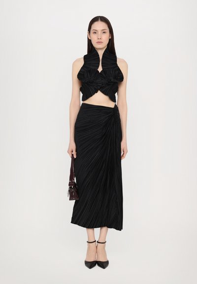 Black pleated crop top with ruffled neckline and matching long skirt. The outfit features a twisting detail on the skirt and sleek shoes.