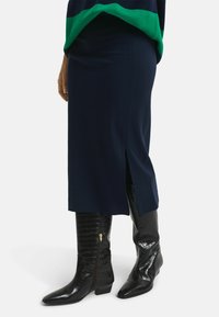 Marineblaue Strickrock mit seitlichem Schlitz, kombiniert mit schwarzen strukturierten Lederstiefeln bis zum Knie, die einen seitlichen Reißverschluss und eine spitze Zehenform aufweisen.