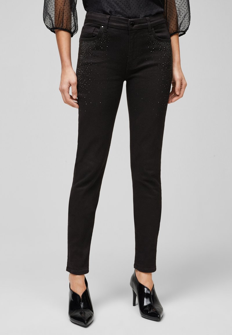 Femme portant un pantalon noir slim avec des strass près des poches et des chaussures à talons hauts pointues noires, debout sur un fond uni.