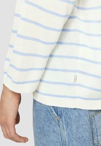 Pull en maille blanche avec des rayures horizontales bleues, présentant un ourlet et des poignets côtelés. Étiquette visible au bord inférieur, porté avec un jean en denim bleu.