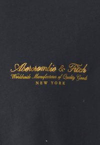 Logo Abercrombie & Fitch haftowane złotem na ciemnym tle, z hasłami "Ogólnokrajowy Producent Towarów Wysokiej Jakości" oraz "Nowy Jork."