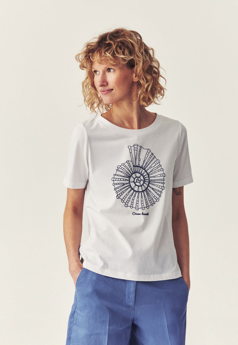 Femme aux cheveux bouclés portant un T-shirt blanc avec un motif de coquille noire en spirale et un pantalon bleu, les mains dans les poches, regardant sur le côté.