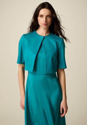 Donna con capelli lunghi e scuri che indossa una giacca a maniche corte color teal sopra un vestito abbinato, in piedi davanti a uno sfondo beige uniforme.