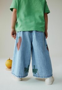 Grøn kortærmet skjorte kombineret med vidde, blå denimbukser med store røde frugt- og grønne bladprint, båret med hvide sneakers.