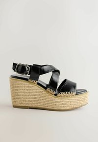 Sandale compensée en cuir noir avec des brides croisées, une fermeture à boucle argentée et une plateforme en paille tissée.