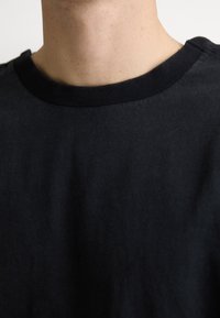 Gros plan sur le cou et la partie supérieure de la poitrine d'une personne portant un t-shirt noir uni à col rond, sur un fond neutre.