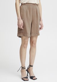 Shorts cargo marron avec une coupe décontractée, fabriqués en tissu léger, dotés de poches latérales et d'accents cousus. Assortis à des sandales à talons noires.