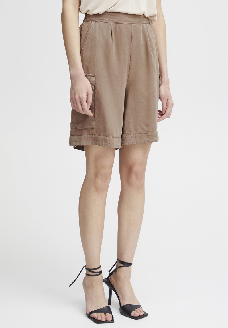 Shorts cargo marron avec une coupe décontractée, fabriqués en tissu léger, dotés de poches latérales et d'accents cousus. Assortis à des sandales à talons noires.