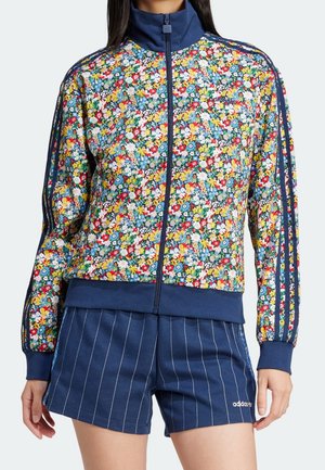 Femme portant une veste zippée à imprimé fleuri avec des poignets bleu marine et un short rayé bleu marine avec le logo Adidas sur la jambe gauche.