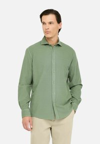Camisa verde de manga larga con cuello button-down, que presenta una tela texturizada y botones beige, combinada con pantalones de color claro.