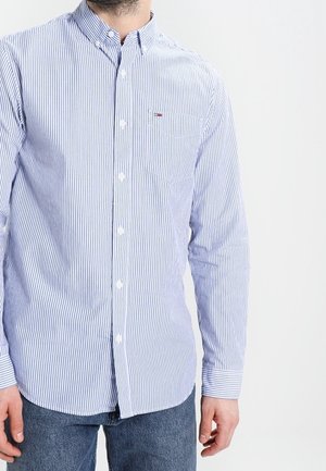 Mand iført en lyseblå og hvid lodret stribet, knap-op skjorte med lange ærmer og mørkeblå jeans.