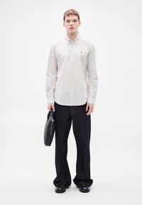 Ριγέ πουκάμισο με κουμπιά σε ανοιχτό μπεζ, συνδυασμένο με χαλαρά σκούρα τζιν. Το μοντέλο κρατά μαύρη τσάντα με φερμουάρ. Τα παπούτσια είναι μαύρα.