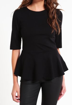 Vrouw draagt een nauwsluitende zwarte top met mouwen tot aan de elleboog en een wijde peplum taille, gecombineerd met zwarte broek.
