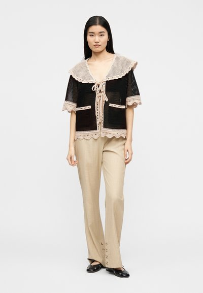 Νεαρή γυναίκα που φοράει μαύρο πλεκτό τοπ με κοντά μανίκια και μπεζ περιθώριο, καθώς και μπεζ παντελόνι με φαρδιά πόδια και λεπτομέρειες κουμπιών στο τελείωμα.