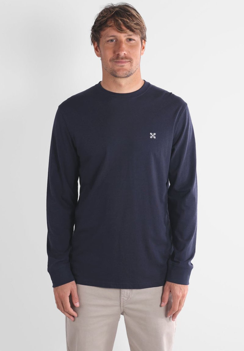 Chemise à manches longues marine en coton doux, dotée d'un col rond et d'un petit logo blanc sur la poitrine, associée à un pantalon de couleur claire.