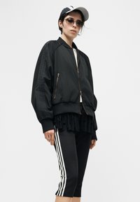 Γυναίκα που φορά μαύρο μπουφάν τύπου bomber, μαύρο παντελόνι cropped με λευκές ρίγες, μαύρη φούστα με βολάν, γυαλιά ηλίου και μαύρο και άσπρο καπέλο μπέιζμπολ.