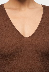 Κοντινό πλάνο ενός ατόμου που φοράει καφέ τοπ με υφή και V-λαιμόκοψη, παρουσιάζοντας την περιοχή του λαιμού και του άνω στήθους.