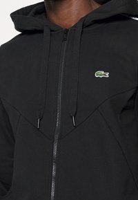 Svart zip-up hoodie i slätt tyg, med en huva, texturerade dragsko och en liten grön krokodillogo på bröstet.