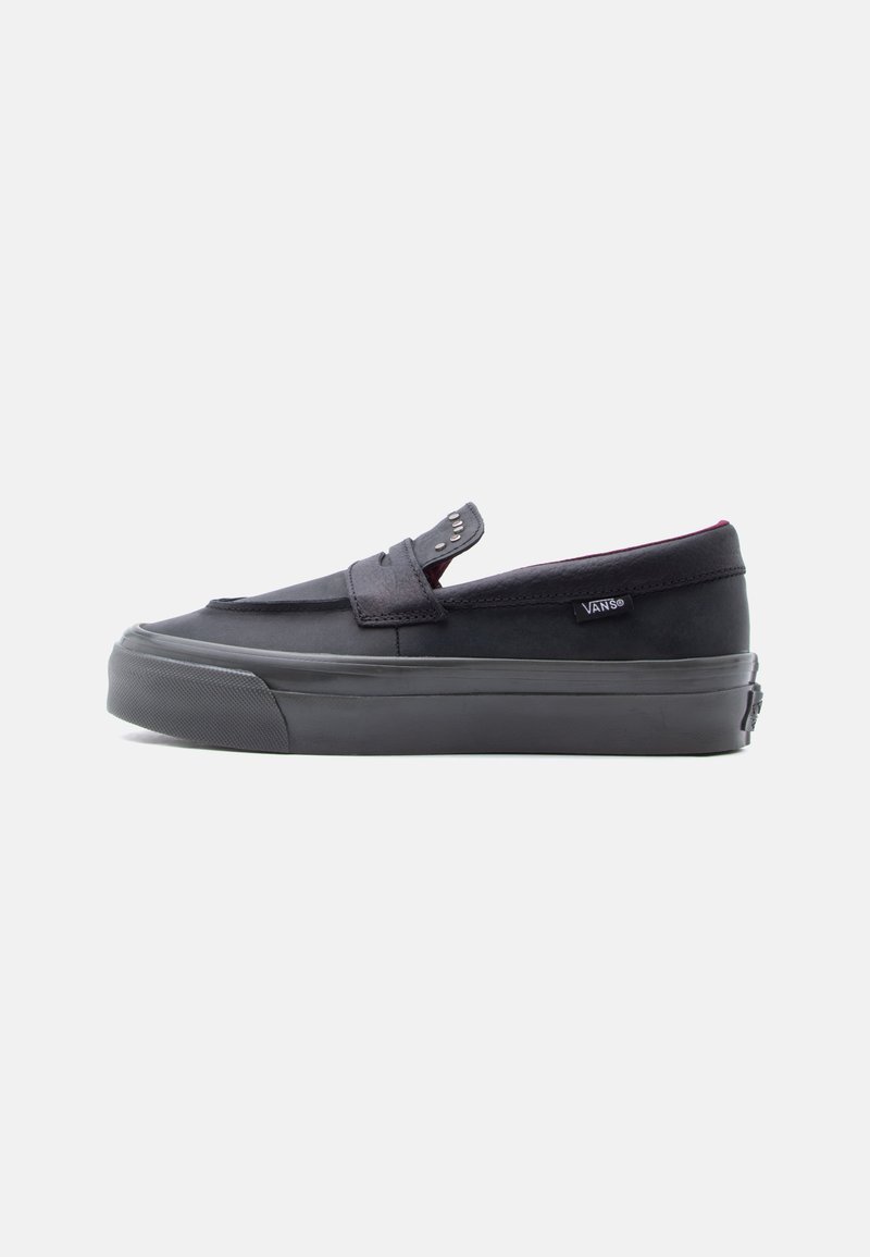 Μαύρο slip-on παπούτσι Vans με λαστιχένια σόλα και διακοσμητικό λουρί, εμφανίζεται σε πλευρική όψη σε λευκό φόντο.