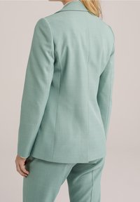 Blazer ajustado verde menta con textura suave, hombros estructurados y abertura única en la parte posterior, combinado con pantalones a juego.
