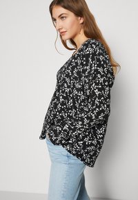 Svart blomstret bluse med lange ermer, v-hals og løs passform. Designet har hvite blomster mønstre på et teksturert stoff.