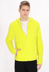 Felpa con zip di colore giallo fluorescente, dotata di tasca a marsupio, texture morbida e cappuccio. Indossata sopra una maglietta bianca e abbinata a pantaloni neri.